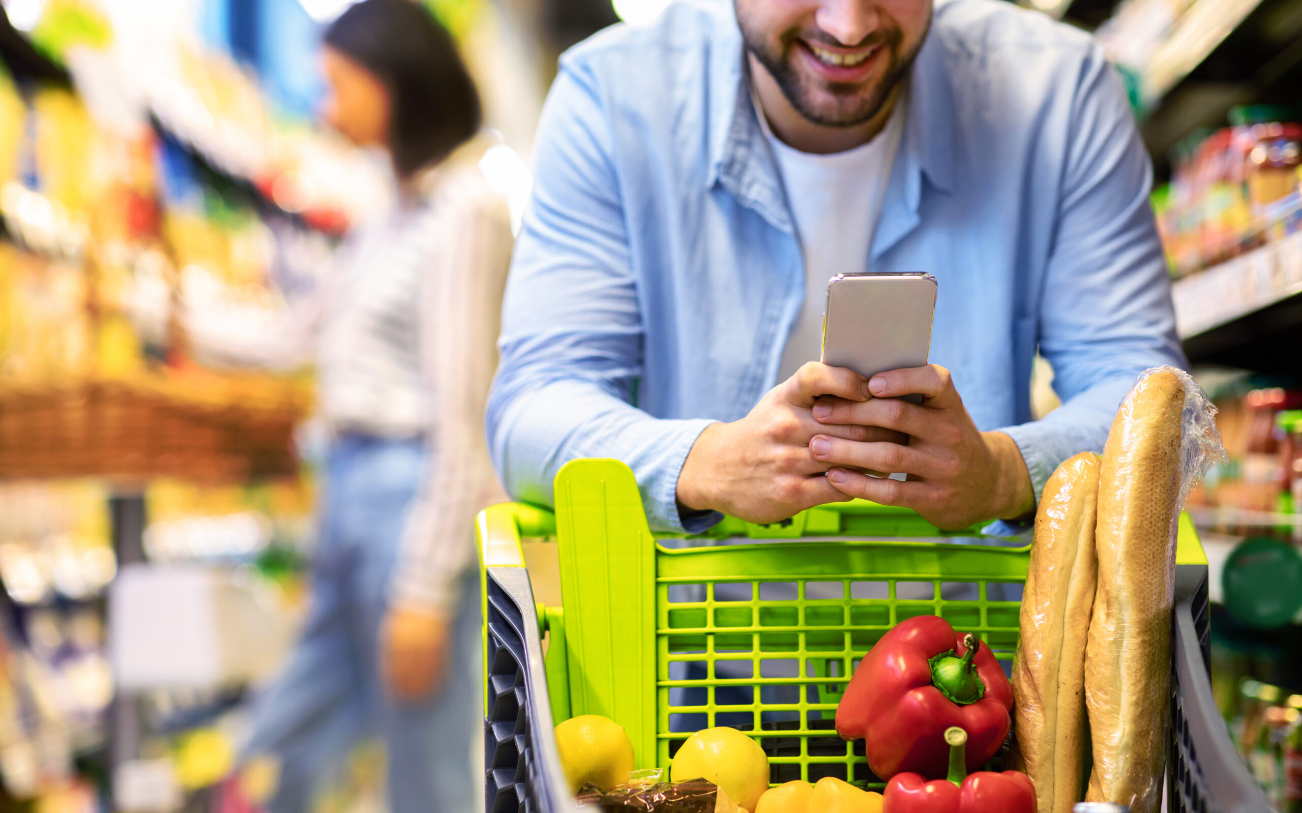 Closeup Of Unrecognizable Man Using Mobile Coupon App For Groceries Shopping Buying Food In Supermarket, Standing With Cart In Hypermarket. Smiling Guy Using Smartphone Purchasing Grocery In Shop